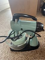 Side and front view of vintage teal Sunbeam mixer showing the attached beaters, the glass mixing bowl, and ceramic bowl inside the glass bowl on the black base.