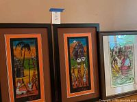 Two framed Batik artworks signed MBIE with orange mats and black frames featuring stylized figures and huts.