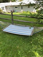 Hammock laid out in green grass, showing blue and gray multicolor striped fabric, wooden spreader bars on each end, and attached to a metal frame stand outdoors.