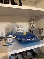 Shelf with various Judaic decor items including cup with Star of David, blue ceramic plates, children's book, porcelain sculpture, and silver tone goblet.