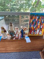 Photo shows four wooden doll figurines wearing colorful traditional fabric dresses standing on a wooden shelf next to a colorful painting by Laurent Casimir depicting a market scene with many abstract figures in bright colors.