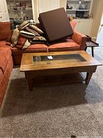 Full front view of rustic wooden coffee table with glass inset top, rectangular shape, and lower shelf, placed in living room with orange couch.