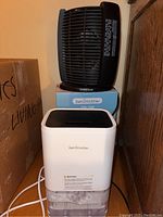 Side view of the white SN-190 dehumidifier with water tank and black Honeywell space heater on top of box behind it.