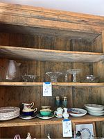 Wooden shelf displaying stacked dinner plates, tea cups and saucers, two decorative wine glasses, creamers and small dishes arranged on shelves