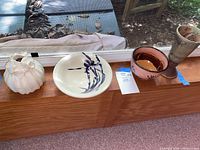 Four ceramic vessels placed on a wooden window sill including a cream vase, a blue floral serving bowl, a pink bowl, and a tall brown vase.