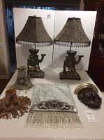 Two camel metal table lamps with patterned fabric lampshades and beaded fringe, two small decorative lampshades, a silver-gray beaded table runner, and a decorative metal sconce arranged on white background.