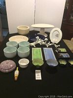 Full lot on black table, showing blue and green sushi plates, light blue bowls, white pedestal dishes, pie plates, purple sea urchin box, and kitchen items