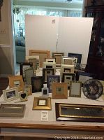 Photo of 27 items comprising 25 picture frames arranged standing and laying, and 2 mirrors. Variety of frame sizes and materials visible.