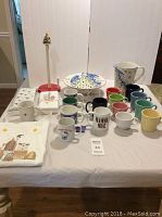 Wide photo of Starbucks Christmas bowl with chip, ceramic pitcher, 8 Fiesta Ware mugs, assorted theme mugs, apron, napkins, and tissue paper on table