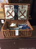 Wicker picnic basket with plates, cutlery secured on lid, and interior showing glasses, napkins, and insulated container.