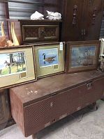 Four framed pictures placed on a brown metal chest with other furniture around. Prints include wildlife and outdoor scenes. Frames show wear and vintage style.