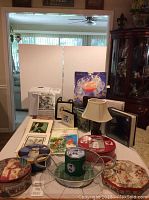 Wide view of lot items laid out on table showing bread maker box, punch bowl set packaging, tins, lamp, stationery, and albums