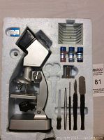 Photo of vintage metal microscope in foam packaging with accessories including tweezers, slides, and small containers.