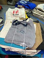 Pile of assorted canvas and burlap bags including a white tote with 'Milbrook' logo and a gray canvas bag with 'Bourne End' embroidery