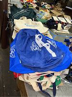 Stack of assorted bags including blue duffel bags and cream canvas tote bags on table