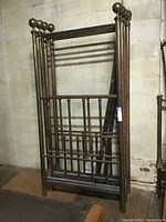 Two twin brass canopy bed frames standing against a brick wall. Both frames include headboards, footboards, and side rails, with vertical rods and round finials on top.