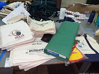 Canvas bags stacked and a bolt of green fabric on a table with various warehouse items