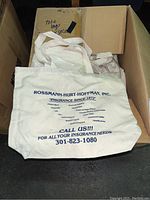Box filled with white canvas tote bags displaying blue printed insurance advertising text.