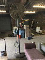 Full view of industrial pedestal fan in warehouse environment showing the base, pole, fan head, and surroundings.