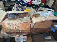 Two cardboard boxes on a wooden surface among other items, one slightly open showing plastic bags and another partially taped closed.