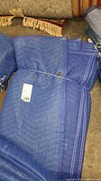 Six cushioned blue moving blankets stacked with visible white stitching and quilted pattern, some stains seen