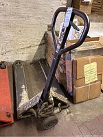 Hydraulic hand pallet truck with rounded metal handle and long lifting prongs, placed next to stacked boxes.