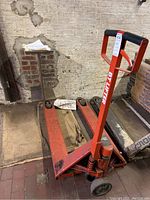 Front angle view of the red hydraulic hand pallet truck with 'BT LIFTER' branding on the handle, showing the handle, forks, and wheels with obvious wear and paint marks.