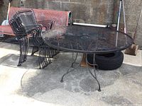 Wrought iron round patio table with a mesh top and stack of eight matching wrought iron chairs next to it, displayed outdoors on concrete.