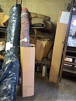 Rolled large area rugs, one wrapped in plastic packaging, standing vertically in storage area.