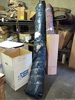 Large rolled rug tightly wrapped in black plastic standing upright with other stored items.