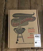 Cardboard box labeled 'The Urban Smokehouse' featuring barbecue imagery and text describing it as a mini-smokehouse to turn backyard barbecue grills into smokers.