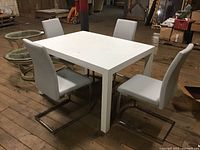 White rectangular table with four matching light gray padded chairs with metal legs in an industrial space. Table shows surface scratches.