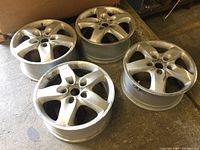 Four matching silver metal tire rims resting on the ground, showing overall condition and wear.