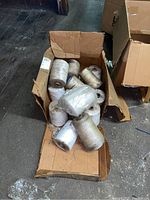 Box containing about a dozen large spools of twine in white and beige tones on a wooden floor.