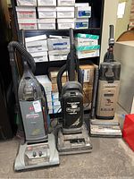 Three Hoover upright vacuums clustered next to each other: one self propelled model on left, black WindTunnel model center, gray WindTunnel on right