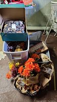Box and plastic bins containing silk flowers, artificial pumpkins, and several bags with autumn decor items grouped in basement setting.