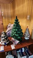 Three ceramic Christmas trees displayed on a table with various background items, showing full lot setup.