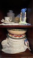 Two shelves of dinnerware including bowls, plates, saucers, mugs, stacked in cabinets with a decorative glass bowl, pitcher, and salt and pepper shakers