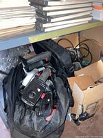 View of multiple black tool cases, a power tool partially visible inside, cords and cardboard boxes on a shelf.