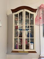 Front view of wooden wall display cabinet with glass-paned double doors, showing interior shelves and figurines inside for scale only.