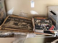Newspaper broadsheets with 'Man on the Moon' headline and other vintage periodicals stacked on a surface.