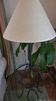 Single table lamp with beige fabric shade and dark metal scrollwork base on glass table near sofa.