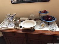 Display of lot items on wooden dresser including stemware, crystal bowl, serving tray, clock, pottery vase, tin fruit stand with apples, and wooden bowl.