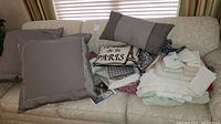 Pile of pillows, folded blankets, and stacked table linens on a cream couch under a window with blinds.