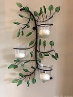 Full view of one candle sconce with three glass holders and decorative green leaf metal branches, mounted on wall and holding white candles.