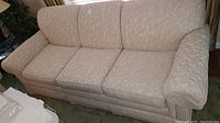 Front angle of beige patterned three seat Thomasville sofa showing textured fabric in embossed floral pattern, cushioned arms and seats.