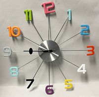Front view of 19-inch retro wall clock with colorful numbers attached on metal rods extending from a circular brushed metal center. Clock hands black with oval detail.