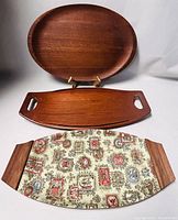 Three mid-century serving trays arranged with oval tray on stand and others on flat surface. Shows size, shape, wood grain, and fabric detail.