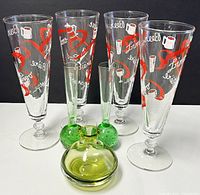 Four vintage pilsner beer glasses and three green glass vases displayed on white surface against black background, showing the glasses' graphics and vase details.