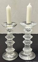 Pair of clear textured glass candleholders with white candles, standing upright on a black surface against a white background.